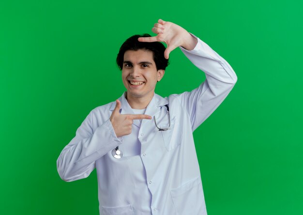 Sonriente joven médico vistiendo bata médica y un estetoscopio haciendo gesto de marco aislado en la pared verde con espacio de copia