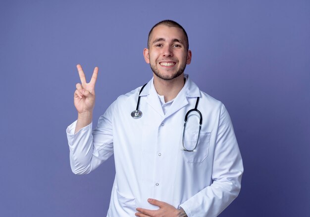 Sonriente joven médico vistiendo una bata médica y un estetoscopio haciendo el signo de la paz y poniendo la mano en el vientre aislado en la pared púrpura
