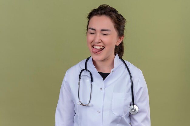 Sonriente joven médico vistiendo bata médica con estetoscopio morder la lengua en la pared verde