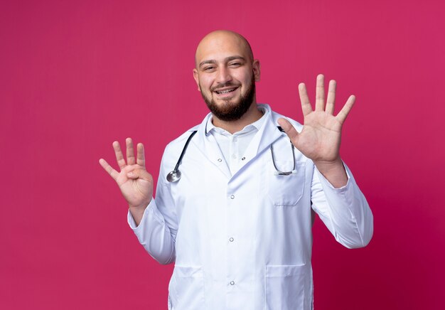 Sonriente joven médico vistiendo bata médica y estetoscopio mostrando diferentes números aislados en rosa