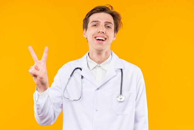 Sonriente joven médico vistiendo bata médica con estetoscopio mostrando gesto de paz aislado en la pared naranja