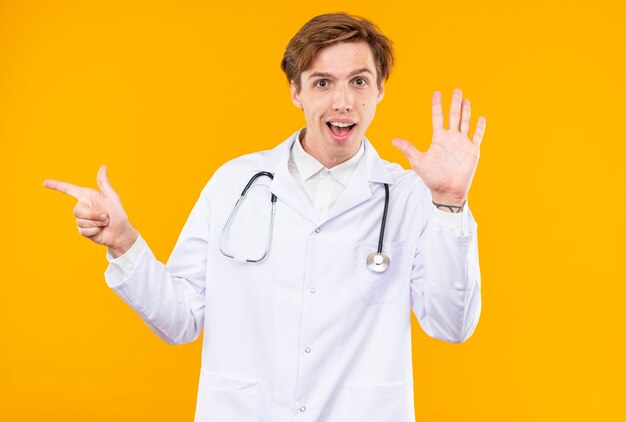 Sonriente joven médico vistiendo bata médica con estetoscopio mostrando puntos de gesto de saludo al lado