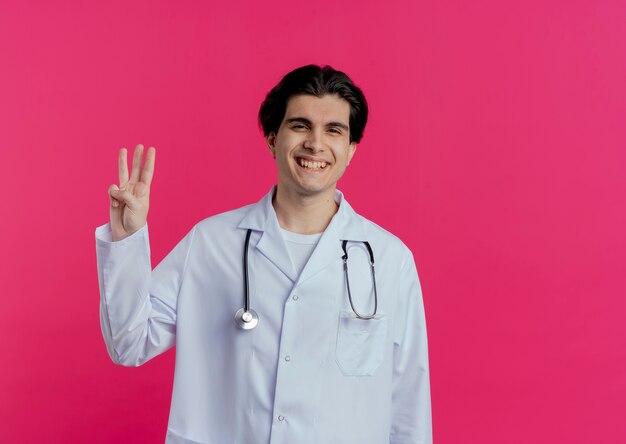 Sonriente joven médico vistiendo bata médica y un estetoscopio mostrando tres con la mano aislada en la pared rosa con espacio de copia