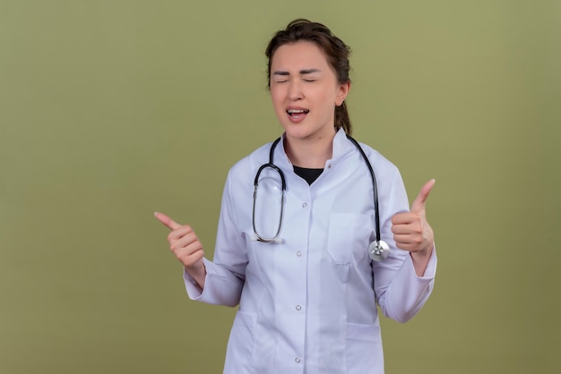 Sonriente joven médico vistiendo una bata médica con estetoscopio ojos cerrados y su tumb sobre pared verde