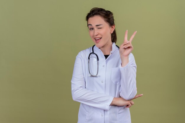 Sonriente joven médico vistiendo bata médica con estetoscopio parpadeando y muestra gesto de paz en la pared verde
