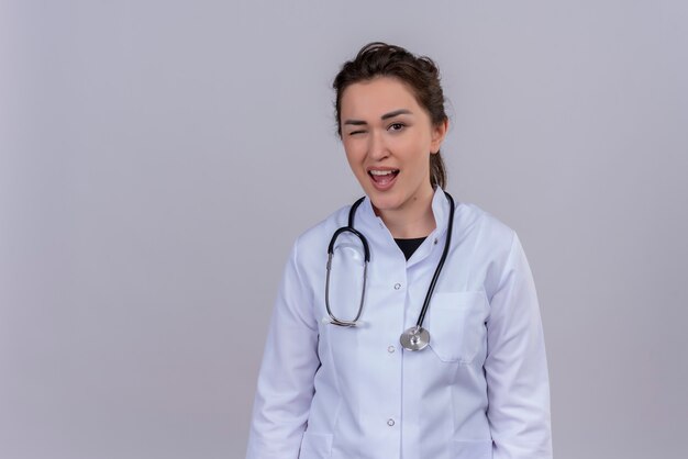 Sonriente joven médico vistiendo bata médica con estetoscopio parpadeó en la pared blanca