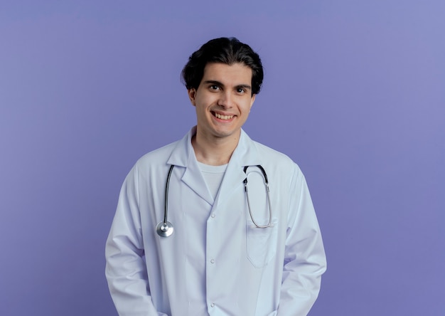 Sonriente joven médico vistiendo bata médica y estetoscopio de pie y aislado en la pared púrpura con espacio de copia