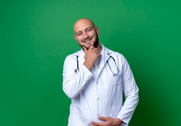 Sonriente joven médico vistiendo bata médica y estetoscopio poniendo la mano en el mentón aislado sobre fondo verde