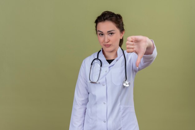 Sonriente joven médico vistiendo bata médica con estetoscopio con el pulgar hacia abajo en la pared verde