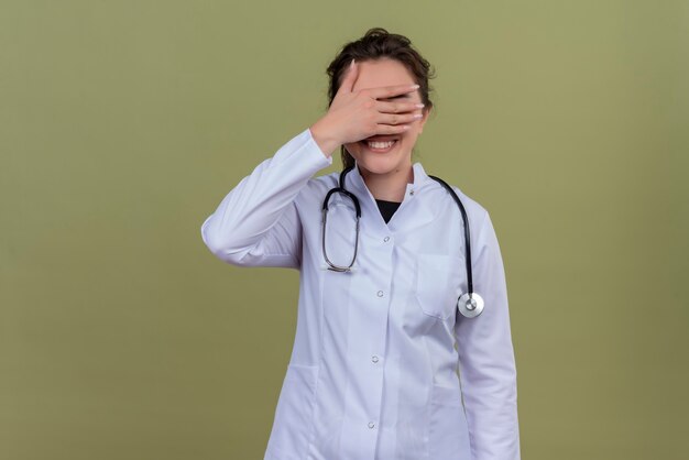 Sonriente joven médico vistiendo bata médica con estetoscopio rostro cubierto con la mano en la pared verde
