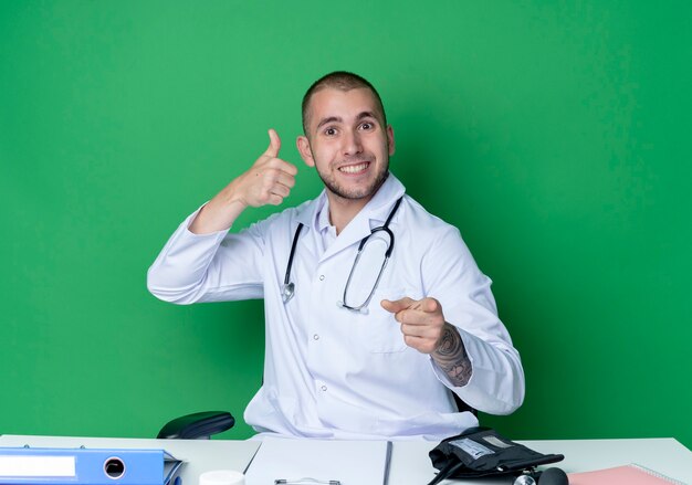 Sonriente joven médico vistiendo bata médica y estetoscopio sentado en el escritorio con herramientas de trabajo mostrando el pulgar hacia arriba y apuntando al frente aislado en la pared verde