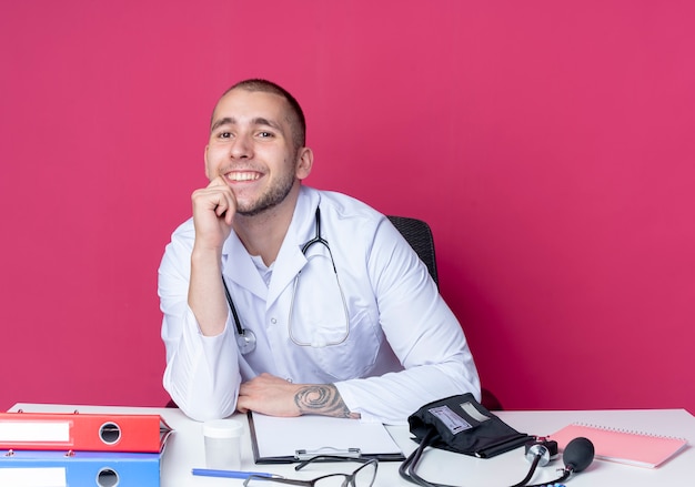 Sonriente joven médico vistiendo bata médica y estetoscopio sentado en el escritorio con herramientas de trabajo poniendo la mano en la barbilla aislada en la pared rosa