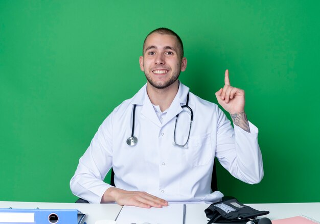Sonriente joven médico vistiendo bata médica y estetoscopio sentado en el escritorio con herramientas de trabajo poniendo la mano sobre el escritorio y levantando el dedo aislado en la pared verde
