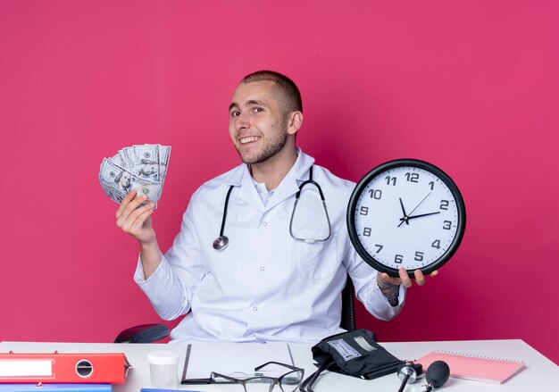 Sonriente joven médico vistiendo bata médica y estetoscopio sentado en el escritorio con herramientas de trabajo con reloj y dinero aislado en la pared rosa