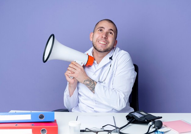 Sonriente joven médico vistiendo bata médica y estetoscopio sentado en el escritorio con herramientas de trabajo sosteniendo el altavoz y mirando hacia arriba aislado en la pared púrpura