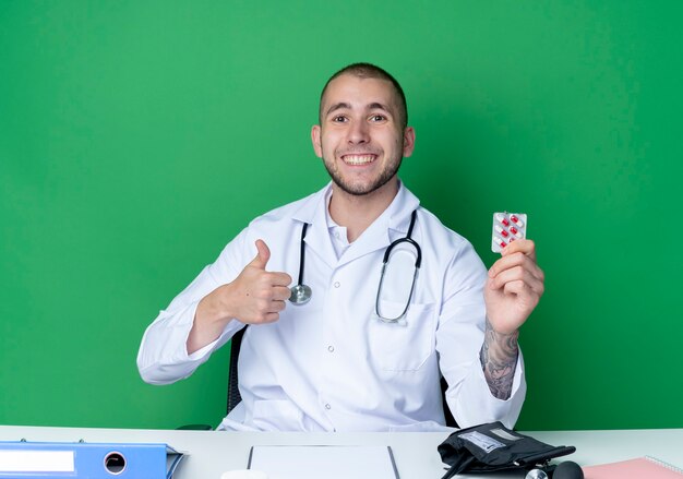 Sonriente joven médico vistiendo bata médica y estetoscopio sentado en el escritorio con herramientas de trabajo sosteniendo el paquete de cápsulas y mostrando el pulgar hacia arriba aislado en la pared verde