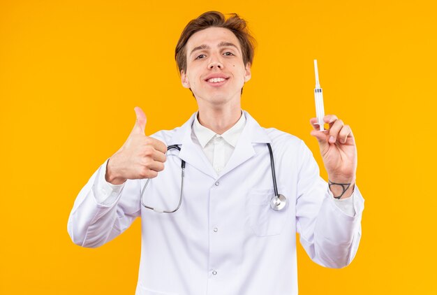 Sonriente joven médico vistiendo bata médica con estetoscopio sosteniendo la jeringa mostrando el pulgar hacia arriba