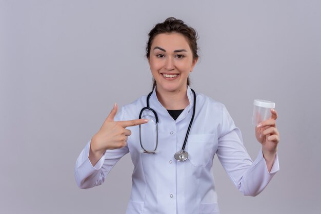 Sonriente joven médico vistiendo bata médica con estetoscopio sosteniendo lata vacía y apunta a lata en la pared blanca