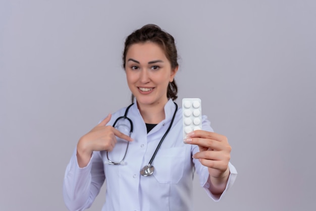 Sonriente joven médico vistiendo bata médica con estetoscopio sosteniendo pastillas y apunta a sí mismo en la pared blanca