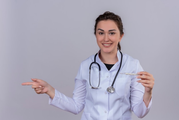 Sonriente joven médico vistiendo bata médica con estetoscopio sosteniendo el termómetro y apunta al lado de la pared blanca