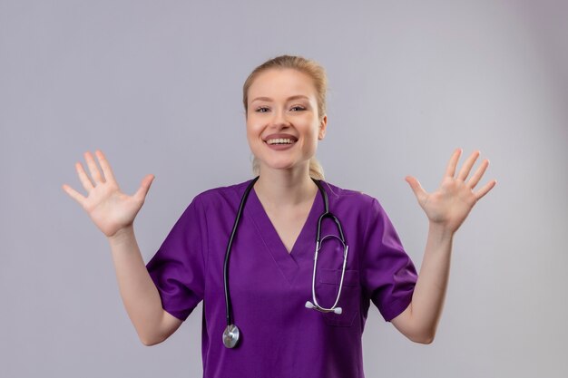 Sonriente joven médico vistiendo una bata médica púrpura y un estetoscopio extiende las manos sobre la pared blanca aislada