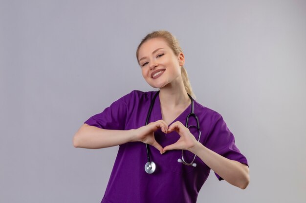 Sonriente joven médico vistiendo una bata médica púrpura y un estetoscopio muestra el gesto del corazón en la pared blanca aislada