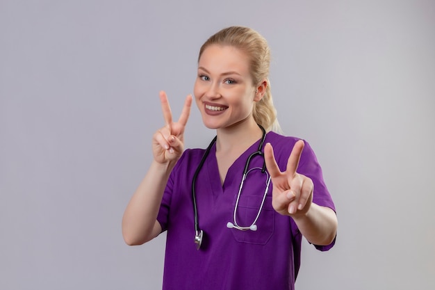 Sonriente joven médico vistiendo una bata médica púrpura y un estetoscopio muestra un gesto de paz con ambas manos en la pared blanca aislada