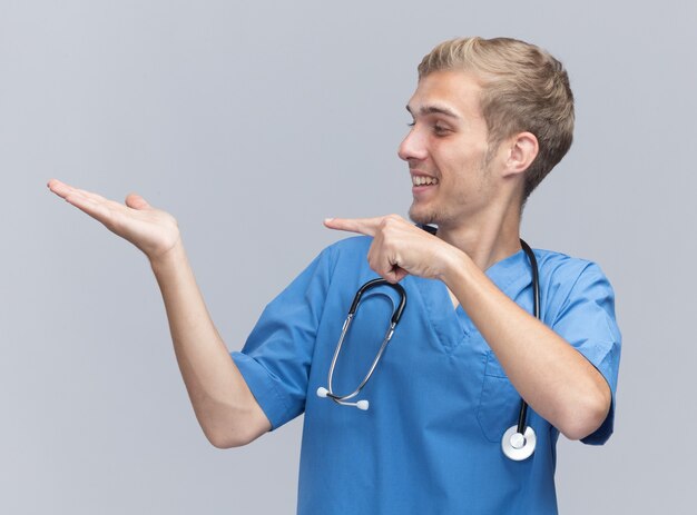 Sonriente joven médico vistiendo uniforme médico con estetoscopio fingiendo sostener y apunta a algo aislado en la pared blanca con espacio de copia