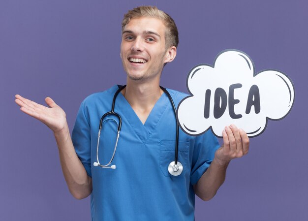 Sonriente joven médico vistiendo uniforme médico con estetoscopio sosteniendo la burbuja de la idea extendiendo la mano aislada en la pared azul