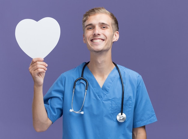 Sonriente joven médico vistiendo uniforme médico con estetoscopio sosteniendo una caja en forma de corazón aislada en la pared azul