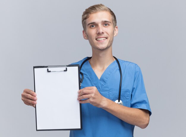 Sonriente joven médico vistiendo uniforme médico con estetoscopio sosteniendo el portapapeles aislado en la pared blanca