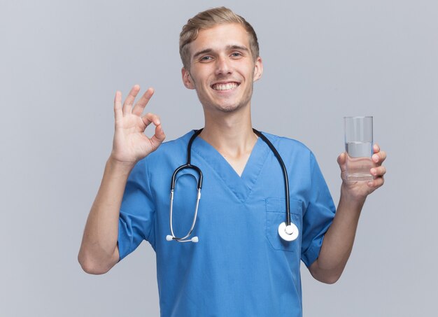 Sonriente joven médico vistiendo uniforme médico con estetoscopio sosteniendo un vaso de agua que muestra un gesto bien aislado en la pared blanca