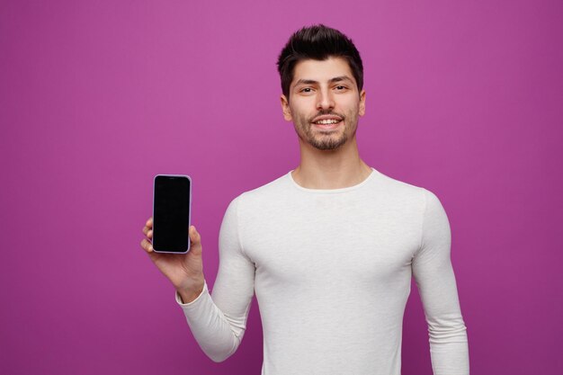 Sonriente joven mirando a la cámara mostrando el teléfono móvil a la cámara aislado sobre fondo púrpura