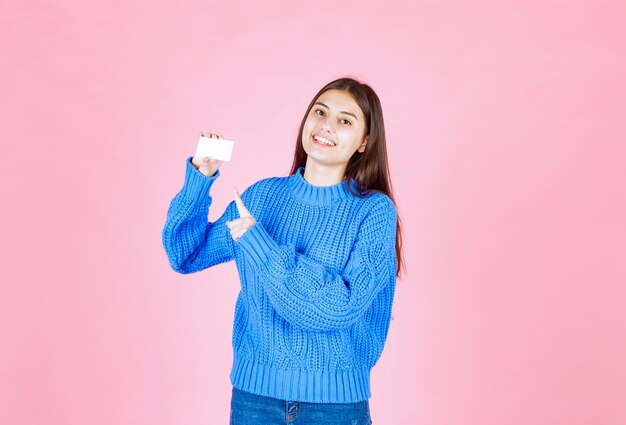 sonriente joven modelo apuntando a una tarjeta en la pared rosa.