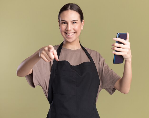 Sonriente joven morena barbero en uniforme apunta a la cámara y sostiene el teléfono aislado en la pared verde oliva con espacio de copia