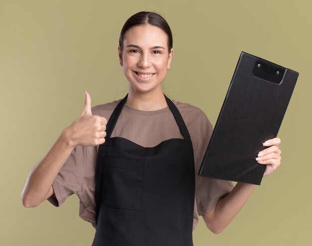 Sonriente joven morena barbero en uniforme pulgar hacia arriba y sostiene el portapapeles