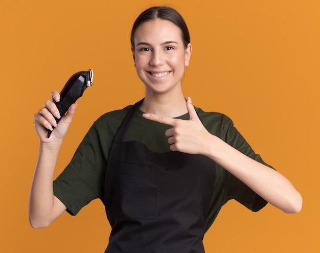 Sonriente joven morena barbero en uniforme sostiene y apunta a cortapelos en naranja