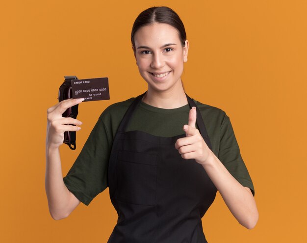 Sonriente joven morena barbero en uniforme sostiene cortapelos y tarjeta de crédito apuntando a la cámara