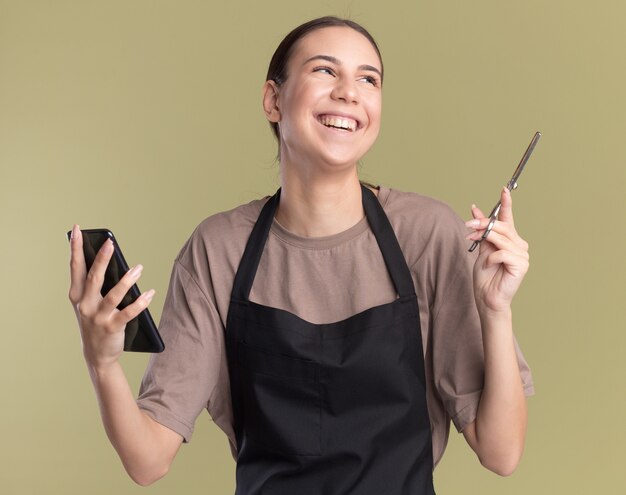 Sonriente joven morena barbero en uniforme sostiene tijeras de adelgazamiento del cabello y teléfono mirando al lado en verde oliva