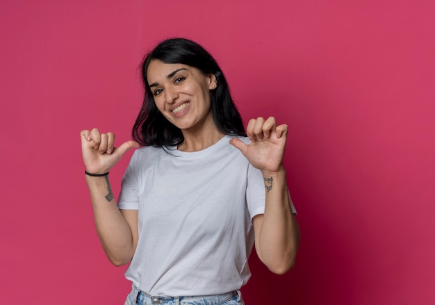 Sonriente joven morena caucásica apunta a sí mismo aislado en la pared rosa