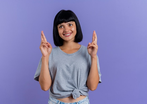 Sonriente joven morena caucásica cruza los dedos mirando al lado aislado sobre fondo púrpura con espacio de copia