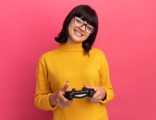 Foto gratuita sonriente joven morena caucásica en gafas ópticas tiene controlador en rosa