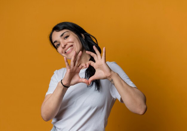 Sonriente joven morena caucásica gestos corazón signo de mano aislado en la pared naranja