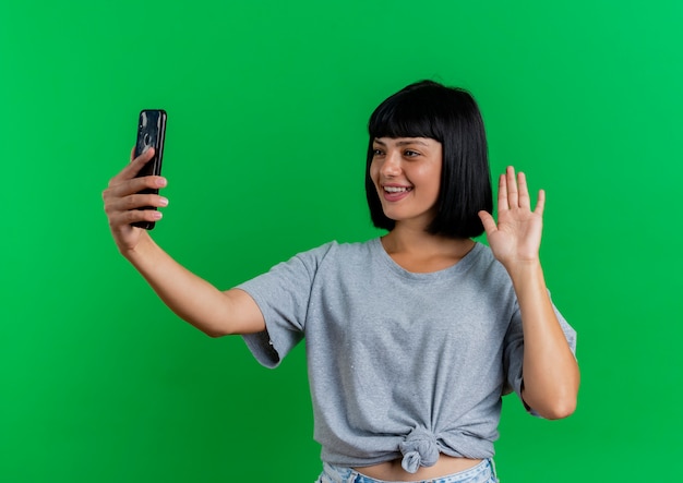 Sonriente joven morena caucásica levanta la mano y mira el teléfono aislado sobre fondo verde con espacio de copia