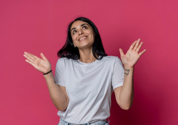 Sonriente joven morena caucásica levanta las manos mirando hacia arriba aislado en la pared rosa