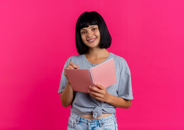 Sonriente joven morena caucásica mira a cámara y sostiene bolígrafo y cuaderno aislado sobre fondo rosa con espacio de copia