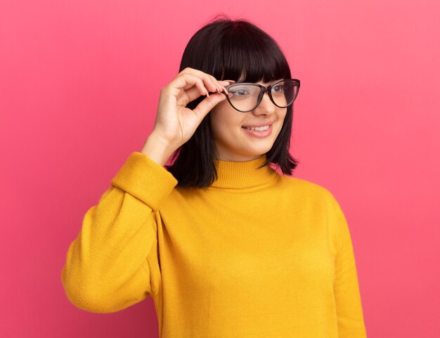 Sonriente joven morena caucásica mira de lado a través de gafas ópticas en rosa