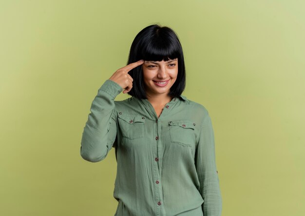 Foto gratuita sonriente joven morena caucásica pone el dedo en la sien mirando al lado aislado sobre fondo verde oliva con espacio de copia