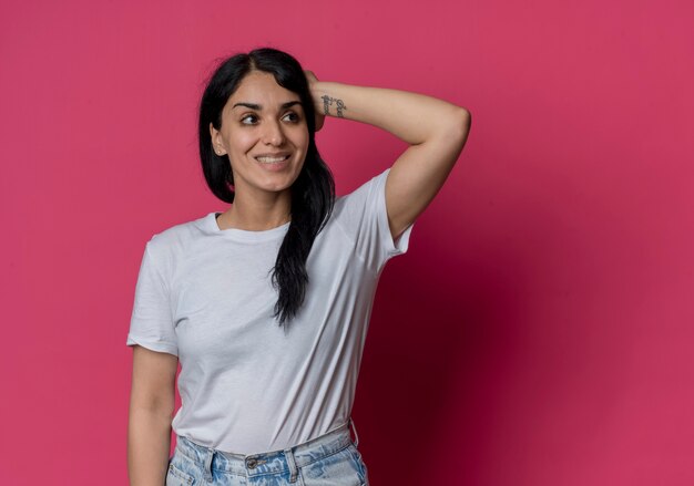 Sonriente joven morena caucásica pone la mano en la cabeza detrás mirando al lado aislado en la pared rosa