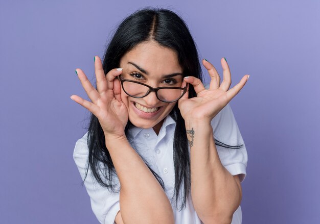 Sonriente joven morena caucásica sostiene gafas ópticas mirando aislado en la pared púrpura